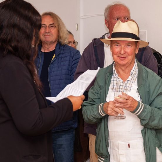 Ein Mann mit Hut hält ein Weinglas, während eine Frau ihm ein Papier gibt. Drei ältere Männer stehen hinter ihnen und beobachten. Sie scheinen sich in einem Raum zu befinden.