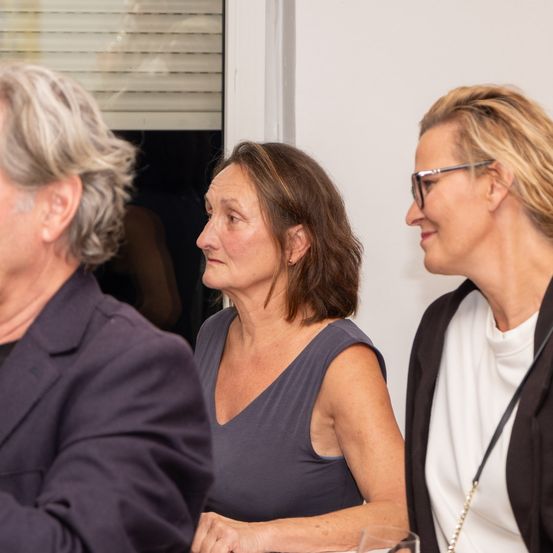 Drei Erwachsene, zwei Frauen und ein Mann, scheinen bei einem gesellschaftlichen Ereignis in ein Gespräch vertieft zu sein, wobei eine Frau eine Brille trägt und eine andere ein Glas in der Hand hält.