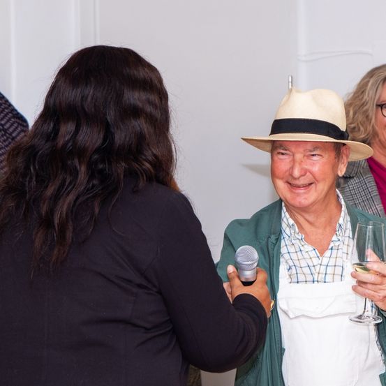 Ein lächelnder älterer Mann mit Hut und Schürze wird von einer Frau interviewt. Er hält ein Weinglas und ein Mikrofon. Eine Frau mit Brille steht hinter ihm.