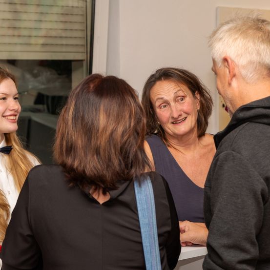 Eine Gruppe von drei Personen unterhält sich in einem Raum. Eine Frau mit lächelndem Gesicht steht in der Mitte. Die Frau auf der linken Seite trägt eine Fliege. Hinter ihnen ist eine weiße Wand.