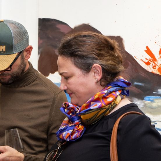 Ein Mann und eine Frau stehen nebeneinander und sehen sich an. Der Mann hält ein Weinglas und die Frau trägt eine Krawatte. Hinter ihnen ist eine Wand mit einem Gemälde.