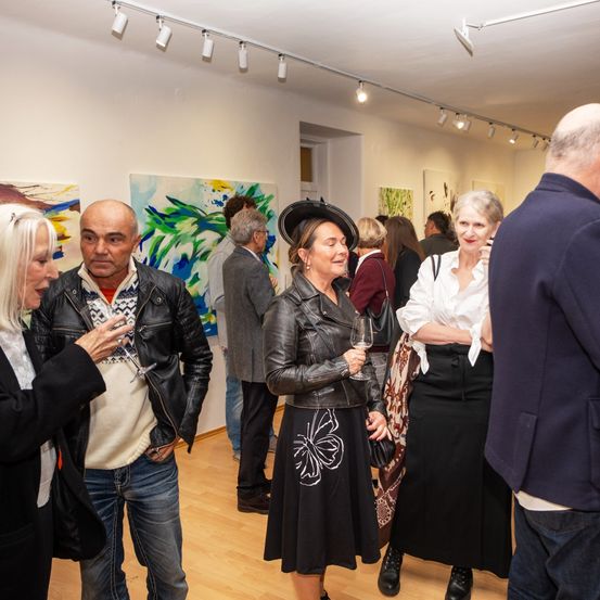 Menschen in einer Kunstgalerie, die Gespräche führen. Eine Frau in Lederjacke und Hut hält ein Weinglas. Bunte abstrakte Gemälde hängen an den Wänden.