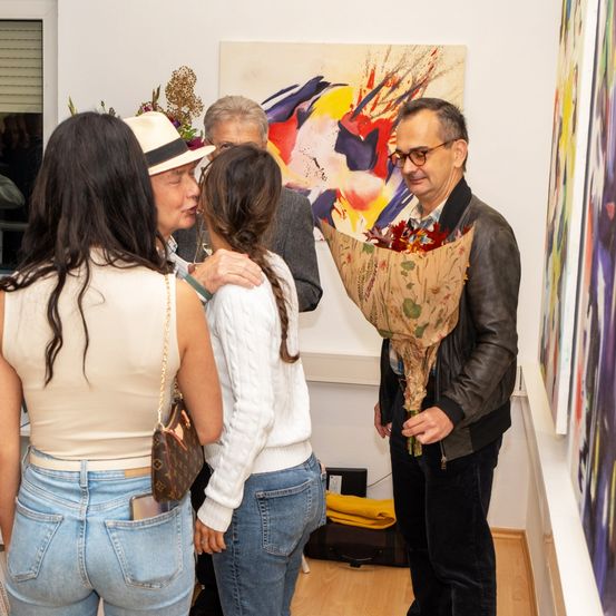 Ein Mann mit Blumen in der Hand steht neben zwei Frauen in einer Kunstgalerie. Dahinter befindet sich ein abstraktes Gemälde an der Wand.