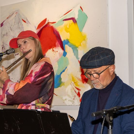 Eine Frau in einem roten Hut spielt eine Flöte und steht vor einem farbenfrohen Gemälde. Ein Mann mit Brille sitzt neben ihr und schaut sie an.