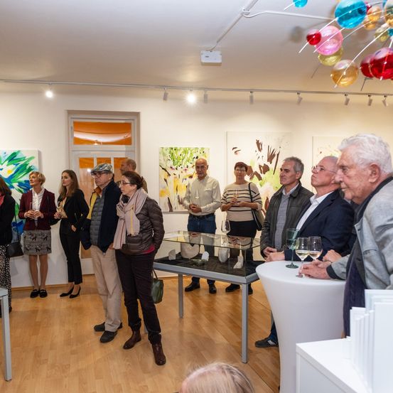 Eine Kunstgalerie mit mehreren Gemälden an der Wand. Menschen stehen herum und betrachten die Gemälde. Es gibt Weingläser und einen Tisch mit einer Glasvitrine im Raum.