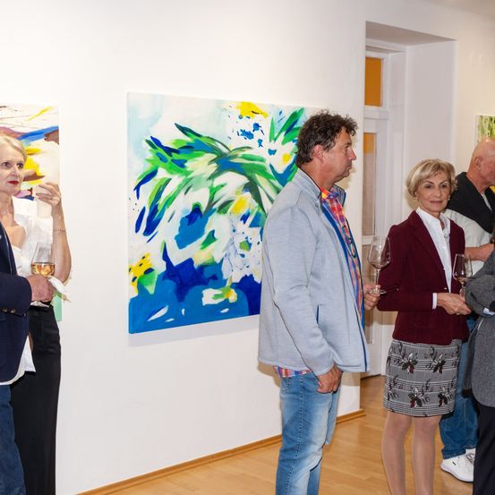 Eine Kunstgalerie mit Menschen, die in einer Gruppe stehen und ein Gemälde betrachten. Sie alle halten ein Weinglas. Die Frau lächelt und sieht das Gemälde an.