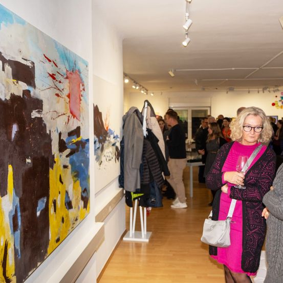 Eine Kunstausstellung, bei der sich Menschen versammeln, um die Kunstwerke zu betrachten. Der Boden ist aus Holz und es gibt Kleidung auf einem Ständer.