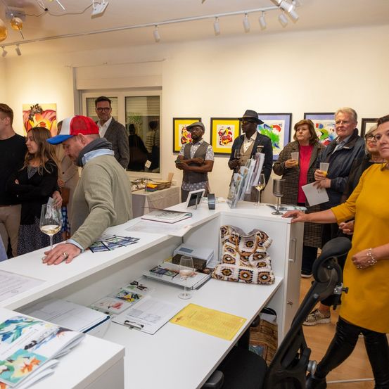 Eine Kunstausstellung mit mehreren Personen um einen Schreibtisch versammelt. Einige halten Weingläser und Papiere. Kunstwerke sind an der Wand ausgestellt.