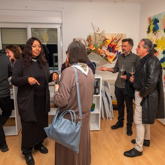 Mehrere Menschen stehen in einer Kunstgalerie und halten Weingläser in der Hand. Eine Frau in Schwarz hält eine Handtasche. Eine andere Frau mit Brille hält ein Glas. Zwei Männer in schwarzen und braunen Ledermänteln unterhalten sich.
