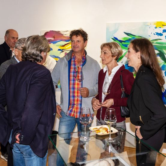 Menschen versammeln sich um einen Glastisch mit Weingläsern und einem Teller mit Essen. Ein älterer Mann hält ein Weinglas. Ein abstraktes Gemälde ist im Hintergrund zu sehen.