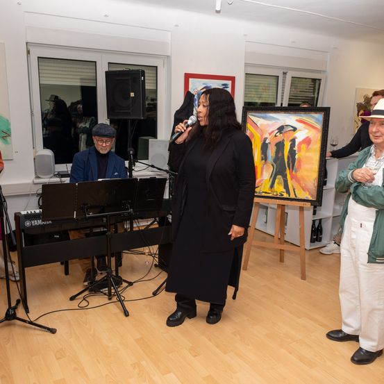 Eine Frau steht vor einem Mikrofon auf der Bühne und singt wahrscheinlich. Ein Pianist spielt hinter ihr. Kunstgemälde hängen an den Wänden, und ein Mann trinkt Wein.