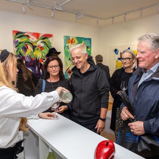 Eine Frau schenkt Wein in ein Glas für einen Mann bei einer Kunstgalerie-Veranstaltung, wo mehrere Leute sich unterhalten und die Kunstwerke betrachten.