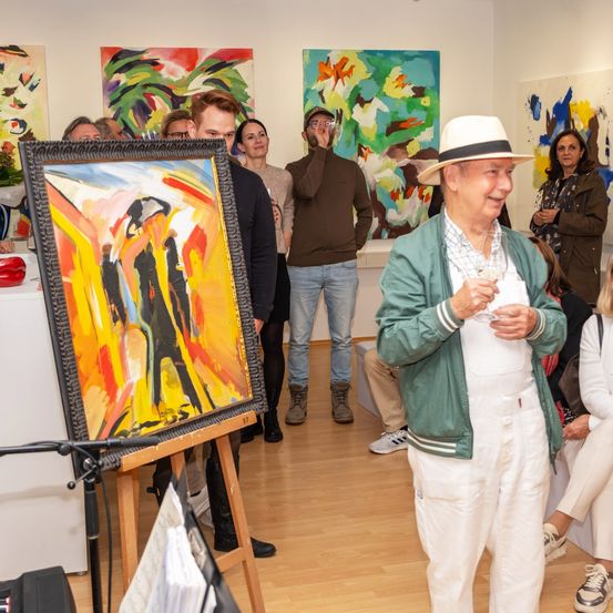 In einer Kunstgalerie betrachten mehrere Personen Gemälde an der Wand. Ein älterer Mann in Hut und grüner Jacke hält ein Glas Wein.