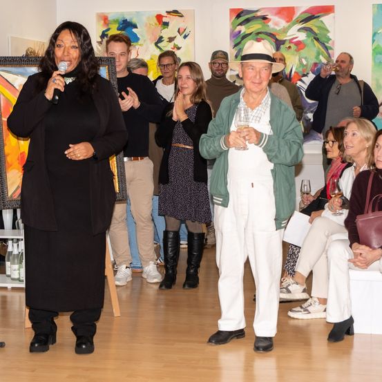 Eine Kunstgalerie mit verschiedenen Gemälden. Eine Frau spricht in ein Mikrofon, während ein Mann ein Glas Wein hält. Menschen sitzen und stehen herum, einige mit Getränken.