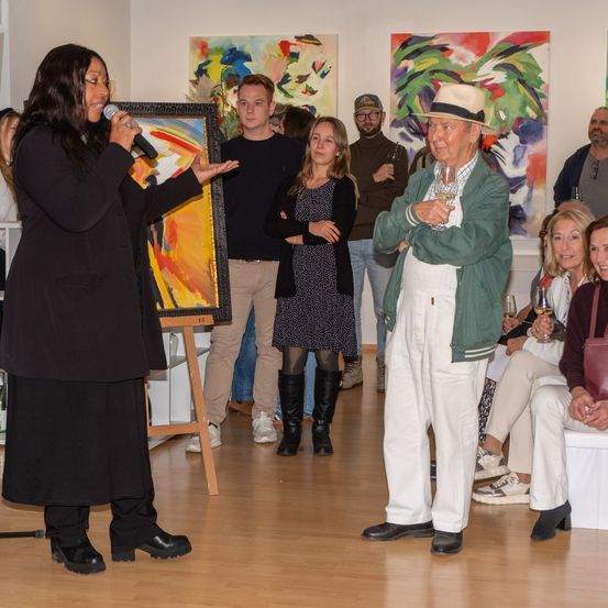 Eine Frau spricht in ein Mikrofon bei einer Kunstgalerie-Veranstaltung. Ein älterer Mann hält ein Weinglas. Leute beobachten die Veranstaltung in einer Kunstgalerie.