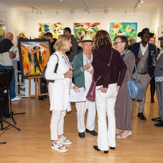 Eine Kunstgalerie mit mehreren bunten Gemälden an den Wänden. Mehrere Menschen stehen und reden, halten Wein glasses. Ein Mann trägt einen Hut.