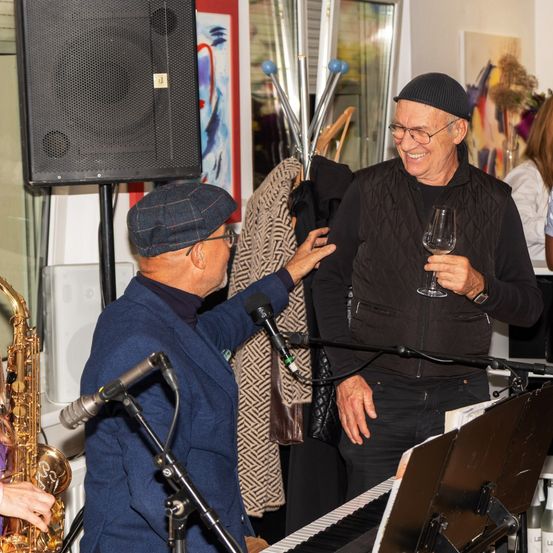 Zwei Männer lächeln und stehen vor einem Mikrofon, einer hält ein Weinglas und der andere spielt die Saxophon. Ein Klavier steht auf der Bühne mit einem Notenständer davor. Dahinter sind ein Lautsprecher und ein Kleiderständer.
