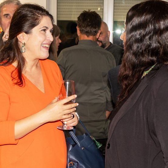 Eine Frau in einem orangefarbenen Kleid, die ein Weinglas hält, spricht mit einer Frau in einem schwarzen Anzug. Hinter ihnen stehen mehrere andere Personen in einem Raum mit einem Glasfenster.