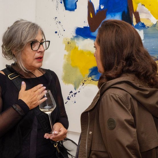 Zwei Frauen unterhalten sich vor einem farbenfrohen abstrakten Gemälde. Die Frau links hält ein Weinglas, während die andere aufmerksam zuhört.