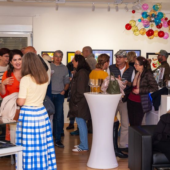 Eine Gruppe von Menschen sozialisiert sich in einer Kunstgalerie. Mehrere Kunstwerke sind an den Wänden ausgestellt. Eine Person hält ein Weinglas.