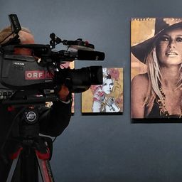 Ein Kameramann mit einer großen Kamera filmt eine Kunstgalerie mit Porträts an der Wand.