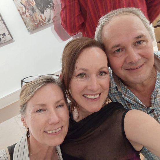 Drei Menschen posieren für ein Selfie in einer Galerie, zwei Frauen lächeln und ein Mann rechts. Hinter ihnen hängen Gemälde an der Wand.