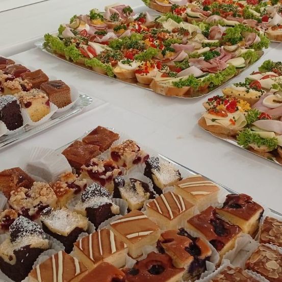 Eine Auswahl an Sandwiches und Desserts ist auf Tabletts ausgestellt. Sandwiches enthalten Fleisch, Tomaten und Salat. Desserts bestehen aus verschiedenen Kuchen und Gebäck mit Toppings.