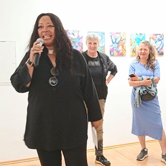 Eine Frau spricht in ein Mikrofon in einer Kunstgalerie, während zwei andere Frauen hinter ihr stehen, eine lächelt und die andere schaut auf ihr Handy. Vier Gemälde hängen an der Wand hinter ihnen.