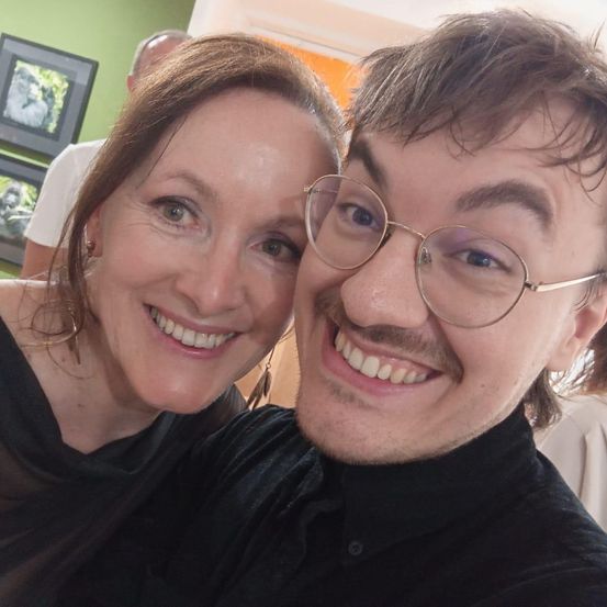 Eine Frau und ein Mann mit Brille lächeln für ein Selfie. Die Frau hat braunes Haar und der Mann einen Schnurrbart. Sie tragen beide schwarze Kleidung. Hinter ihnen ist eine grüne Wand mit Bilderrahmen.