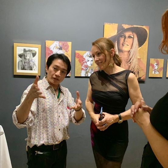 Eine Kunstausstellung mit einer Frau in einem schwarzen Kleid, die einen Stift hält, neben einem lächelnden Mann. Kunstgemälde sind an einer grauen Wand im Hintergrund montiert.