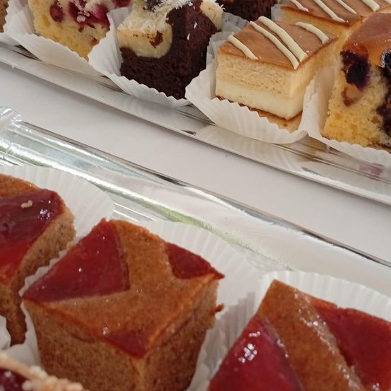 Eine Auswahl an Desserts auf Papierbechern steht auf einem Glastablett. Die Desserts umfassen Kuchen mit roter Füllung, Käsekuchen und einen Schokoladenkuchen mit Kokosnuss.