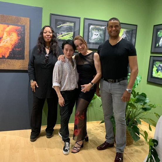 Vier Personen stehen vor einer grünen Wand mit Tiergemälden und lächeln für ein Foto. Zwei Frauen und zwei Männer, einer mit Halskette und Uhr, der andere mit Gürtel.