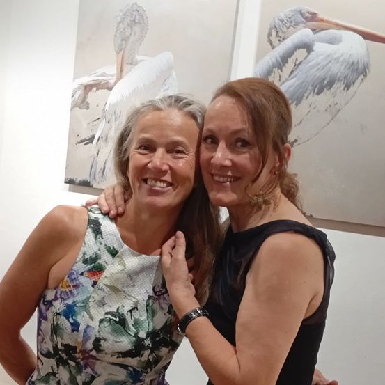 Zwei Frauen stehen lächelnd zusammen vor einer Wand mit Pelikangemälden.
