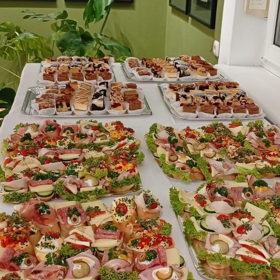 Eine Buffettafel mit Tabletts von Sandwiches, Gebäck und Desserts, vor einer grünen Wand mit Pflanzen und gerahmten Bildern.