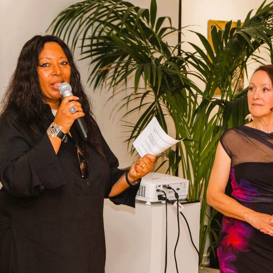 Eine Frau mit langen Haaren spricht in ein Mikrofon und hält ein Papier in der Hand. Eine andere Frau steht in der Nähe, beide sind formell gekleidet.