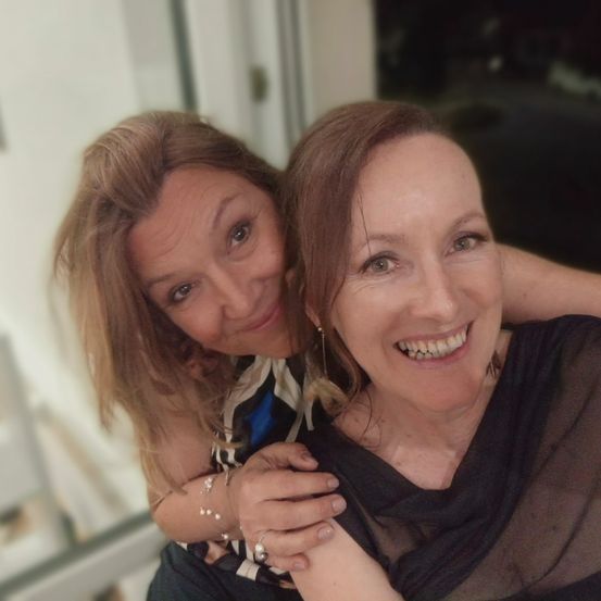 Zwei Frauen umarmen sich, lächeln und posieren für ein Foto. Die Frau links hat blonde Haare und trägt ein Armband, während die Frau rechts braune Haare und eine Halskette hat.