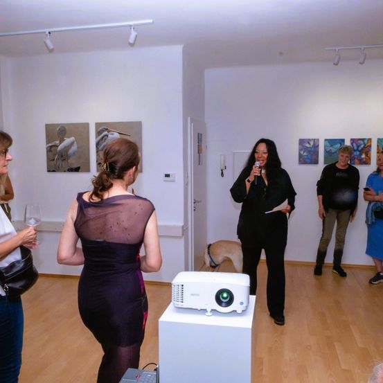 In einer Kunstgalerie spricht eine Frau in ein Mikrofon, während andere sich versammeln, mit Gemälden an den Wänden und einem Projektor ausgestellt.