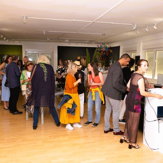 Eine Gruppe von Menschen in einem Raum, wahrscheinlich bei einer Veranstaltung. Einige unterhalten sich, während andere trinken. Der Raum hat Holzböden und weiße Wände.