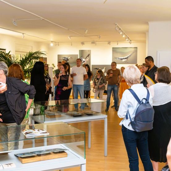 Eine Gruppe von Menschen versammelt sich in einer Kunstgalerie und interagiert mit verschiedenen ausgestellten Kunstwerken. Eine Frau mit Rucksack spricht mit einer anderen Frau.