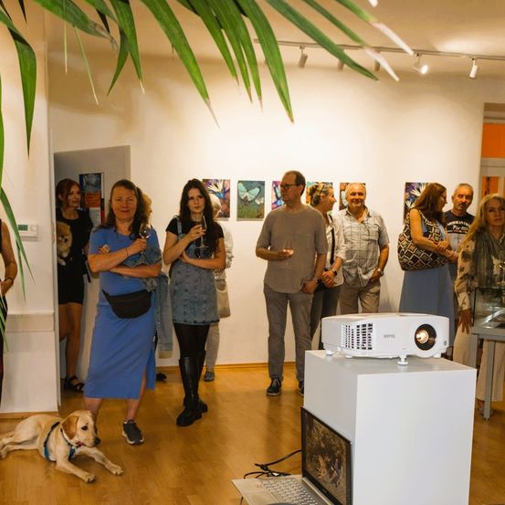 Eine Gruppe von Menschen befindet sich in einer Kunstgalerie. Sie stehen um einen Beamer auf einem Ständer mit einem Laptop herum. Sie trinken Wein und unterhalten sich. Eine Frau trägt ein blaues Kleid, und ein Hund liegt auf dem Boden.