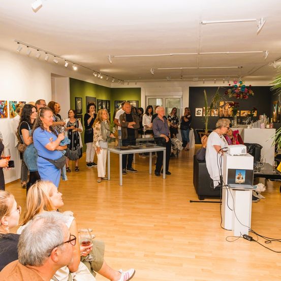 Eine Kunstausstellung mit Menschen in einem Raum. Einige Leute sitzen und andere stehen. Eine Person hält ein Mikrofon und scheint zu sprechen.