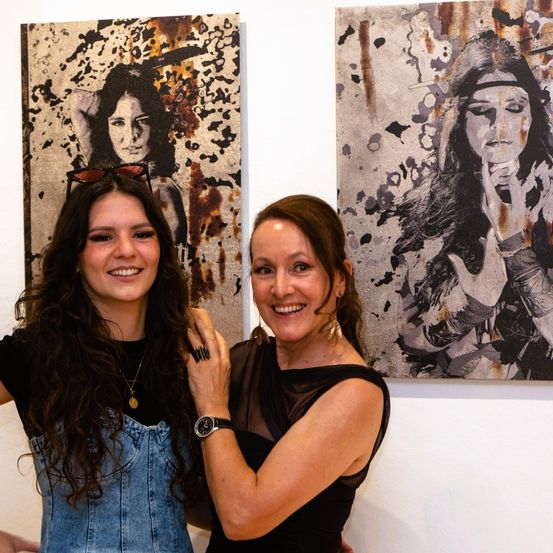 Zwei Frauen posieren neben einer Wand mit zwei Gemälden. Die Frau links trägt eine Overall aus Denim. Die Frau rechts trägt ein schwarzes Oberteil. Beide lächeln.