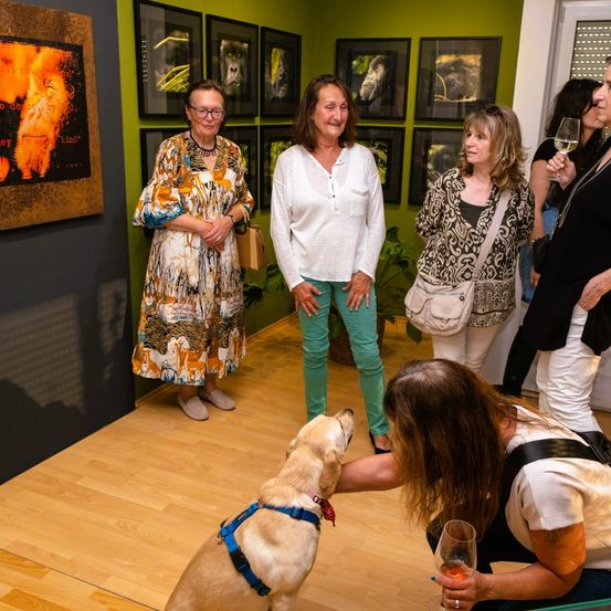 Mehrere Frauen in einer Galerie, eine mit einem Hund, genießen Getränke. Gerahmte Tierfotos an grünen Wänden, eine Frau lehnt sich an eine Pflanze.