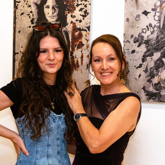Zwei Frauen stehen vor einer Galeriewand mit abstrakter Kunst und lächeln in die Kamera. Eine trägt ein Jeans-Overall, die andere ein schwarzes Kleid. Sie posieren eng beieinander.