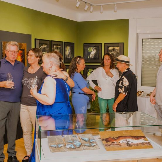 Eine Gruppe Erwachsener in einem Raum mit grünen Wänden und montierten Bildern. Sie betrachten eine Glasvitrine mit Kunstwerken. Der Mann vorne hält ein Weinglas.