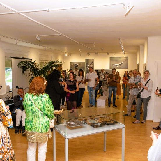 Eine Gruppe von Menschen in einer Galerie betrachtet verschiedene Kunstwerke an der Wand und einen Glastisch mit Objekten.