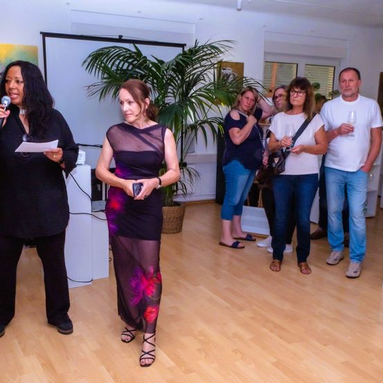 Eine Gruppe Erwachsener steht in einem Raum und hört einer Frau mit Mikrofon zu. Eine Frau in der Mitte hält ein kleines Gerät. Ein Topf mit Pflanzen steht hinter ihnen.