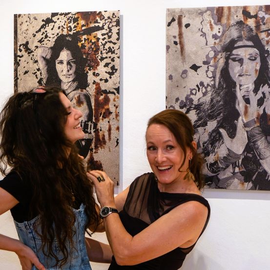 Zwei Frauen lächeln nebeneinander, wobei eine von ihnen ein Gemälde einer Frau mit langen Haaren an einer weißen Wand betrachtet.
