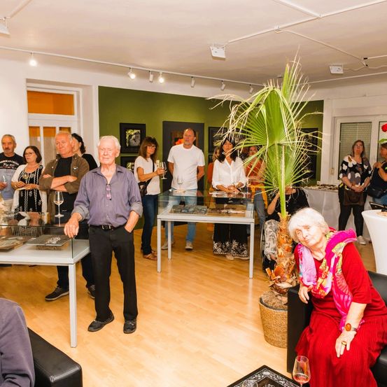 Eine Gruppe von Menschen steht und sitzt in einem Raum mit Holzboden und Wänden. Ein Mann mit weißen Haaren steht mit den Händen in den Taschen. Eine Frau sitzt auf einer Couch. Es gibt Tische mit ausgestellten Gegenständen.