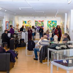 Eine Gruppe von Menschen befindet sich in einer Kunstgalerie, einige sitzen und andere stehen. Ein Pianist spielt in der Ecke. Mehrere Gemälde hängen an den Wänden.